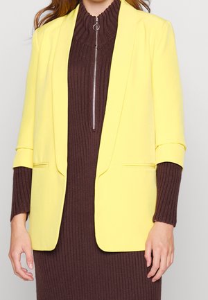 Kvinde iført brun ribbet kjole med lynlås, lagdelt med en lys gul blazer med optrukne ærmer, stående mod en ensfarvet baggrund.