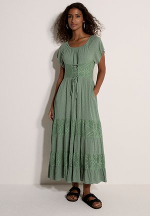 Kobieta ubrana w zieloną sukienkę do kostek z koronkowymi zdobieniami, krótkimi rękawami i czarnymi sandałami, stoi na tle jednolitej jasnej ściany.