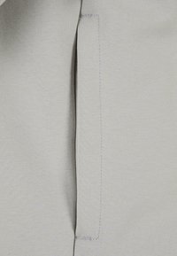 Ljust grått tyg med en slät struktur och subtil ribbing. En sydd fickdetalj är synlig på sidan.