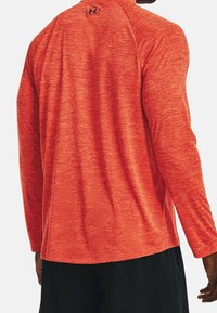 Man wearing red long-sleeve athletic top with black Under Armour logo on upper back and black shorts, standing with back facing forward.