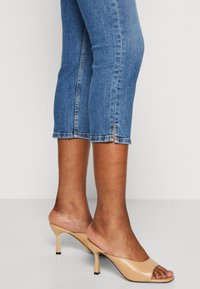 Beige öppna tofflor med en tunn stilettklack. Slät läderdesign med en elegant stil. Matchas med korta blå jeans med sidslitsar.