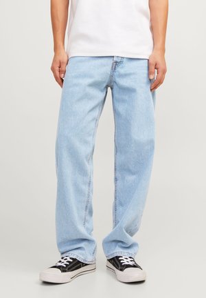 Personne portant un jean ample bleu clair, un t-shirt blanc et des baskets basses noir et blanc, debout devant un fond uni.