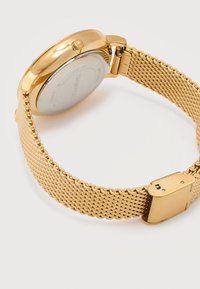 Μεταλλικό μπρασελέ σε χρυσή απόχρωση με κούμπωμα και στρογγυλή πίσω όψη κάσας ρολογιού, όπου εμφανίζονται οι ενδείξεις "Swiss Made" και το λογότυπο Swarovski.