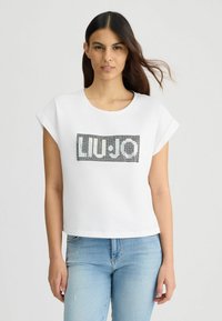 Bílý krátký rukáv crop top vyrobený z měkké tkaniny, s černým a stříbrným flitrovým logem umístěným na přední straně.