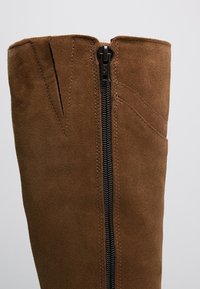 Bota de ante marrón hasta la rodilla con una cremallera negra, que presenta un acabado texturizado y un corte en ángulo en la parte superior para un aspecto estructurado.