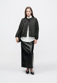 Jachetă neagră matlasată cu nasturi pression, cămașă albă, fustă maxi din piele neagră și pantofi negri. Modelul ține o geantă de umăr, stând în fața unui fundal simplu.