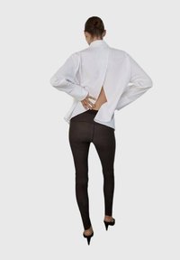 Camicia bianca a maniche lunghe con un taglio verticale sulla schiena, abbinata a leggins marroni scuri e tacchi alti neri. Design minimalista.