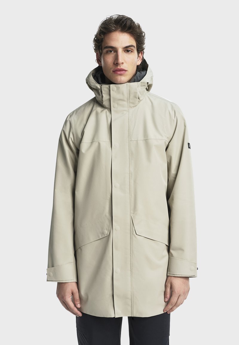 Svijetlosmeđa vodootporna jakna s visokim ovratnikom, kopčanjem na dugmadi i velikim prednjim džepovima. Glatka tekstura i podesiva kapuljača.