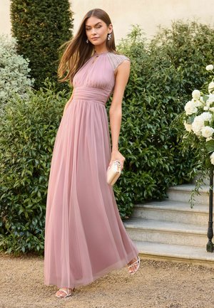 Frau in einem bodenlangen, mauvefarbenen Kleid mit perlenbesetzten Schulterdetails steht im Freien nahe Treppen und Grün, hält eine helle Clutch.