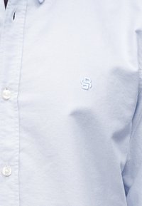 Ανοιχτό μπλε πουκάμισο με κουμπιά, κατασκευασμένο από ύφασμα με υφασμάτινη υφή. Διαθέτει μικρό κεντημένο λογότυπο στο στήθος και κουμπιά που μοιάζουν με πέρλες.