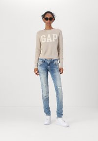 Beige knitted sweater with "GAP" logo, paired with light blue distressed jeans and white sneakers. Accessories include black sunglasses.