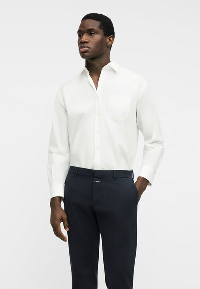 Homme portant une chemise blanche à manches longues avec des boutons et une poche poitrine, ainsi qu'un pantalon sombre, debout avec une main dans la poche.