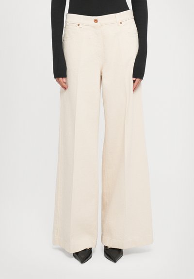 Femme portant un pantalon large taille haute crème, des chaussures noires à bout pointu et un haut noir à manches longues, se tenant contre un fond uni.