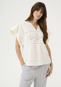 Blusa color crema con scollo a V e maniche corte a sbuffo. Presenta un intricata ricamo a occhiello e delicati dettagli perferati.