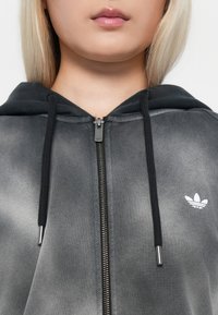 Hooded zip-up sweatshirt in gradient gray fabric, featuring a metallic zipper, black drawstrings, and a white logo on the left chest.