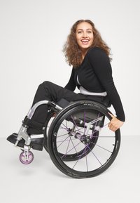 A black and white-clothed person seated in a wheelchair with purple accents, featuring large wheels and a woven backrest, smiling.