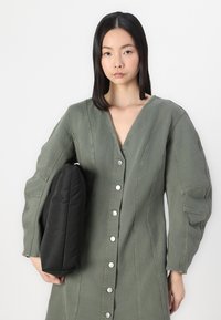 Olivno zelena denim obleka z V-izrezom, dolgimi rokavi in srebrnimi gumbi. Črn oblazinjen dodatek, ki se drži pod pazduho. Gladka tekstura.