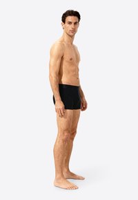 Black fitted swim shorts, made of stretchy material. Flat waistband, smooth texture, and a minimalist design. Model posed sideways.