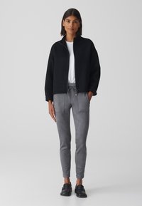 Schwarze Zip-Jacke mit hohem Kragen, kombiniert mit grauen schmalen Jeans mit Fronttaschen. Schwarze glatte Slipper runden das Outfit ab.