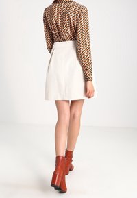 Pitkähihainen paita, jossa on geometrinen kuosi ruskean ja beige värissä, yhdistetty valkoiseen hameeseen ja ruskeisiin korkea-antoisiin nilkkasaappaisiin. Silkkinen kangas.