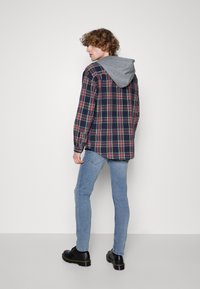 Camisa de franela a cuadros en azul marino, rojo y verde, con capucha gris; combinada con jeans de mezclilla azul claro y zapatos negros. Diseño casual con mangas largas.