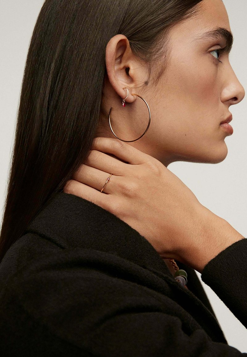 Pendientes de aro en metal dorado, con un pequeño arete con una piedra preciosa roja, llevados con un abrigo negro. Se destacan texturas suaves y elegantes.