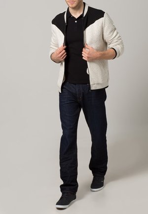 Jasnoszara kurtka zapinana na zamek z teksturowanym wzorem, czarne akcenty na ramionach, czarna koszulka polo, granatowe dżinsy i ciemne sneakersy.