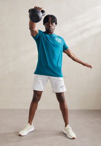 Atlet v tyrkysovom tričku a šedých šortkách, zdvíhajúci čiernu kettlebell. Obuv sú svetlé tenisky. Vnútorné prostredie s neutrálnymi tónmi.