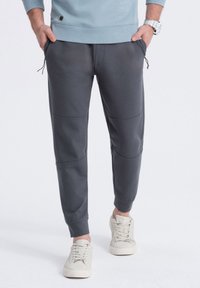 Pantalones de chándal grises con dos bolsillos laterales, puños elásticos y detalles de panel contrastantes. Combinados con zapatillas de sport gris claro.