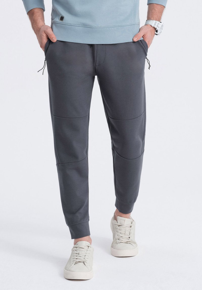 Pantalones de chándal grises con dos bolsillos laterales, puños elásticos y detalles de panel contrastantes. Combinados con zapatillas de sport gris claro.