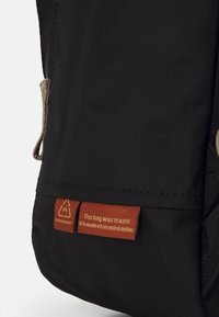 Sac noir en nylon recyclé avec une surface texturée, une poche latérale et deux étiquettes indiquant le matériau et la marque.