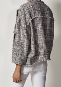 Szary i niebieski, fakturowany żakiet z tweedu o luźnym kroju, postrzępionych krawędziach i rękawach do trzech czwarci, noszony na białych spodniach.