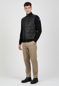 Czarna kamizelka puchowa z wysokim kołnierzem, zamkiem błyskawicznym z przodu oraz bocznymi kieszeniami, noszona na czarnej koszuli z długim rękawem i beżowych spodniach, w zestawieniu z czarnymi butami.