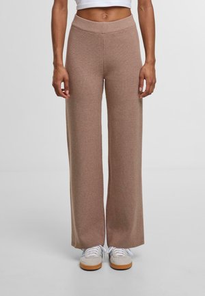 Woman wearing beige textured wide-leg pants and white sneakers, standing against a plain light gray background.