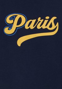 Text galben în stil cursiv "Paris" cu contur brodat albastru pe litera P, așezat pe un fundal de material bleumarin închis.