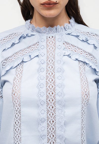 Γυναίκα που φοράει ανοιχτό μπλε μπλούζα με κάθετες πάνελ από δαντέλα με λουλούδια, βολάν δαντέλας στους ώμους και δαντελένιο υψηλό λαιμό με κυματιστό χείλος.