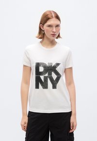 Λευκό T-shirt με μεγάλο μαύρο γκλίτερ λογότυπο "DKNY" στο μπροστινό μέρος. Κοντά μανίκια και κλασικός στρογγυλός λαιμός. Συνδυάζεται με μαύρο παντελόνι cargo.