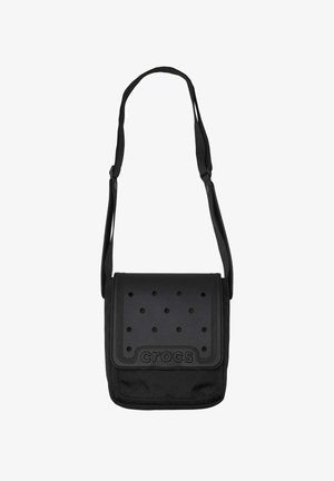Black crossbody bag with a textured rubber flap, circular cutouts, and a fabric body. Features an adjustable strap and embossed logo.