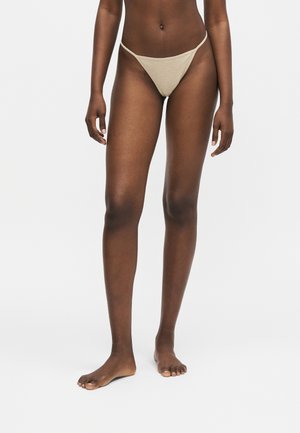 Persona delgada de pie descalza con ropa interior beige de corte alto sobre un fondo claro y sencillo, piernas y torso visibles.