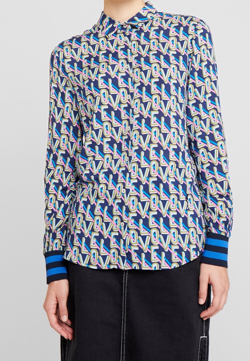 Napapaita, jonka pohjaväri on laivastonsininen ja jossa on värikäs "LOVE"-kuvio. Hihansuut ovat siniraidalliset, paidassa on kaulus ja pitkät hihat.