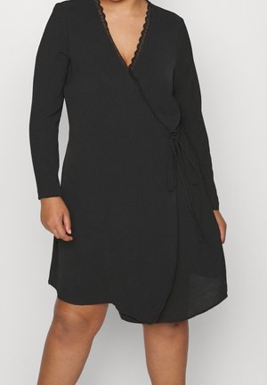 Kobieta w czarnej sukience kopertowej z długim rękawem, ozdobionej koronką przy dekolcie i wiązaniem na boku, stojąca na jednolitym tle.