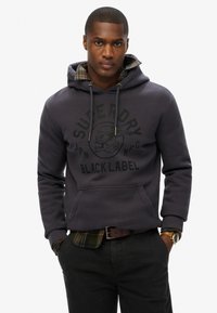 Donkergrijze hoodie met logo-opdruk, voorzak met kangoeroe-sluiting en trekkoordkap. Gelaagd over een groen geruit shirt, gecombineerd met donkere broek.