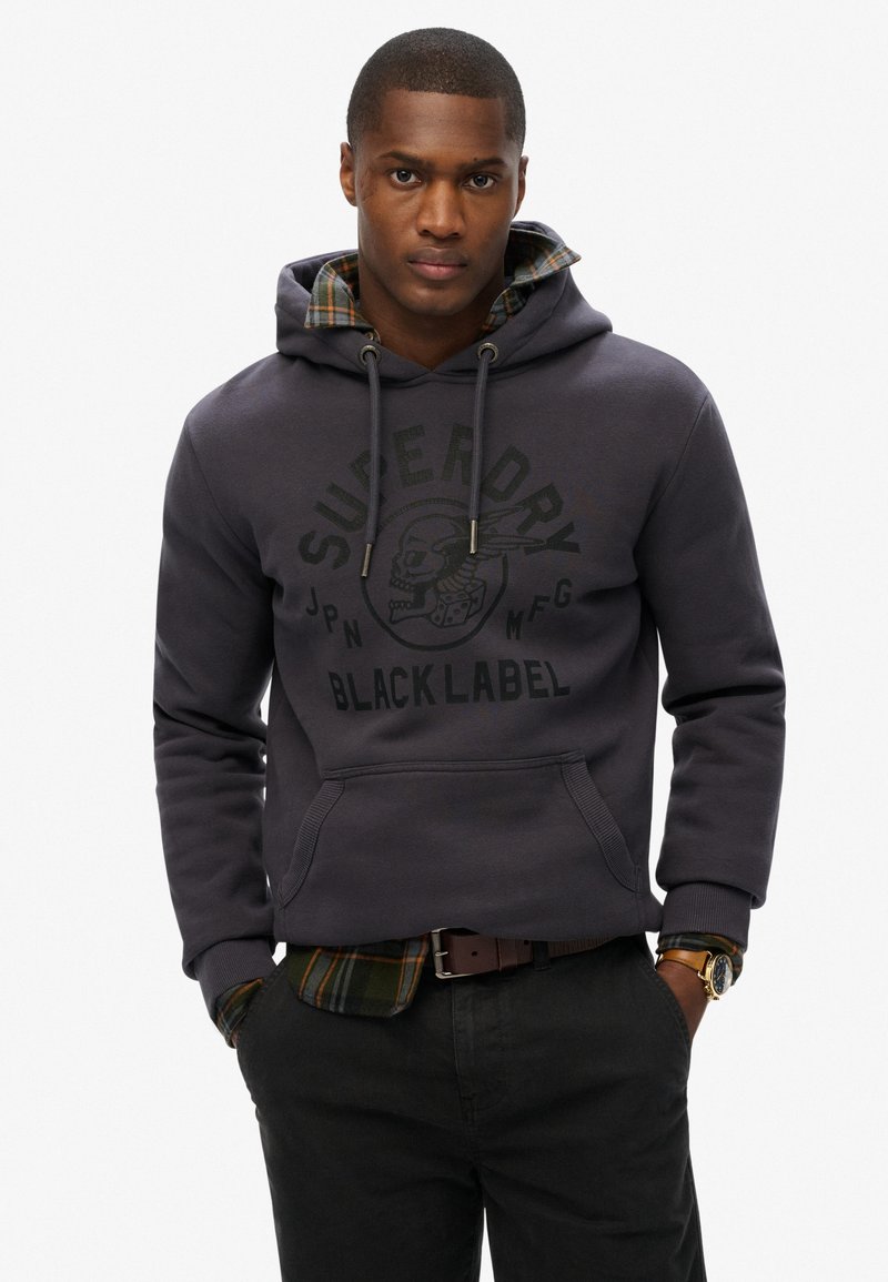 Donkergrijze hoodie met logo-opdruk, voorzak met kangoeroe-sluiting en trekkoordkap. Gelaagd over een groen geruit shirt, gecombineerd met donkere broek.