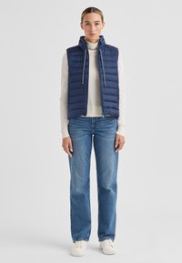 Marineblaue Steppweste mit Kapuze, gesteppter Optik, leichtem Material, Frontreißverschluss, kombiniert mit einem cremefarbenen Rollkragenpullover und blauen Jeans.