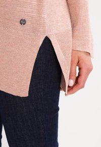 Light pink ribbed sweater with side slits, paired with dark blue denim. Features a small branded metal accent near the hem.