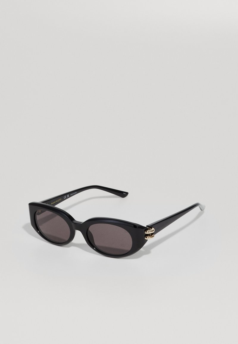 Occhiali da sole neri con una montatura audace e angolare. Presentano lenti scure e dettagli dorati sulle tempie. Texture liscia e finitura lucida.