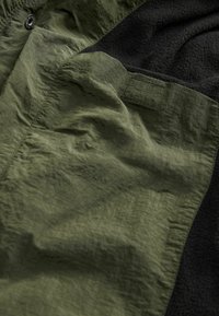 Olivgrön tyg med en rynkad textur, kombinerat med en svart fleece-del. Har en knapp och kontrasterande sömnadsdetaljer.