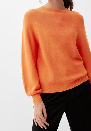 Donna che indossa un maglione arancione lavorato a maglia, largo e a maniche lunghe, abbinato a pantaloni neri su uno sfondo neutro.