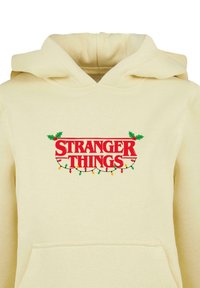 Krém színű kapucnis pulóver kenguru zsebbel, pirossal írva "STRANGER THINGS", zöld fagyöngy és színes karácsonyi fényekkel körülvéve.