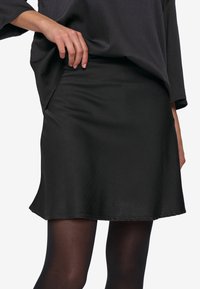 Gonna nera in un tessuto liscio e lucido con un orlo leggermente svasato e una vita aderente. Indossata con una maglietta grigia ampia e collant scuri.